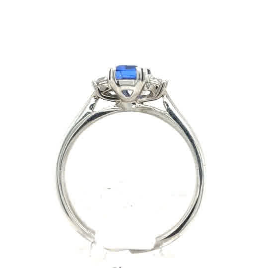 The Tanzanite and Diamond Trilogy Dress Ring in 9ct white gold features a blue rectangular tanzanite gemstone at the center with intricate detailing beneath the setting, shown from the side.
