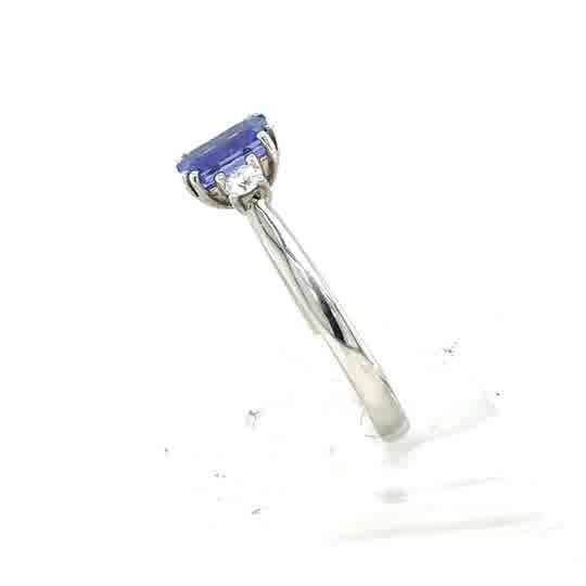 A 9ct white gold Tanzanite and Diamond Trilogy Dress Ring with a rectangular blue tanzanite in four prongs, side view on a white background.
