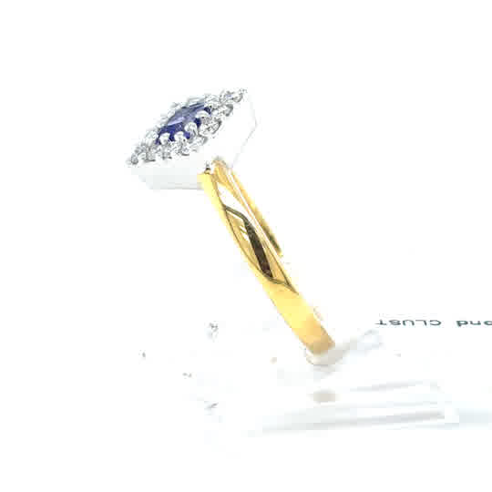 The Tanzanite and Diamond Cluster Dress Ring in 18ct yellow gold showcases a round blue tanzanite center, encircled by sparkling diamonds, displayed on a white stand and viewed from the side.