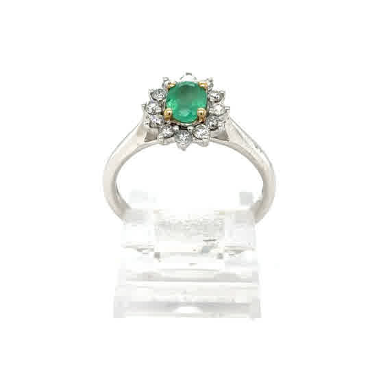 The Emerald and Diamond Cluster Ring in 9ct white and yellow gold features an oval emerald center with a floral halo of sparkling diamonds, elegantly displayed on a clear stand.
