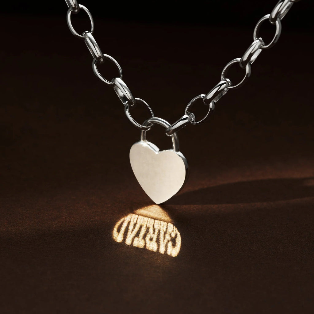 A silver chain necklace with a heart-shaped pendant casts a shadow on a dark surface, reflecting "GRATEFUL" in capitals—a style reminiscent of the Clogau Cariad® Sterling Silver Padlock Bracelet.