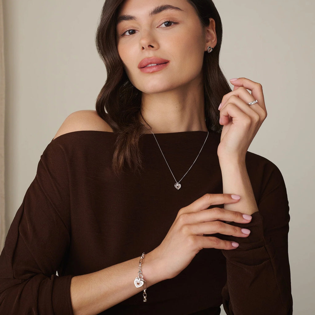 Woman wearing a dark brown off-shoulder top, showcasing the Clogau Cariad® Sterling Silver Sparkle Figaro Bracelet with Topaz, a heart-shaped necklace, earrings, and ring against a plain beige background.