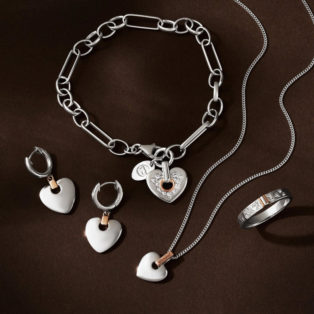 A Clogau Cariad® Sterling Silver Sparkle Figaro Bracelet with Topaz displayed on a brown background, alongside matching heart-shaped earrings, a heart pendant necklace, and a diamond-accented ring.