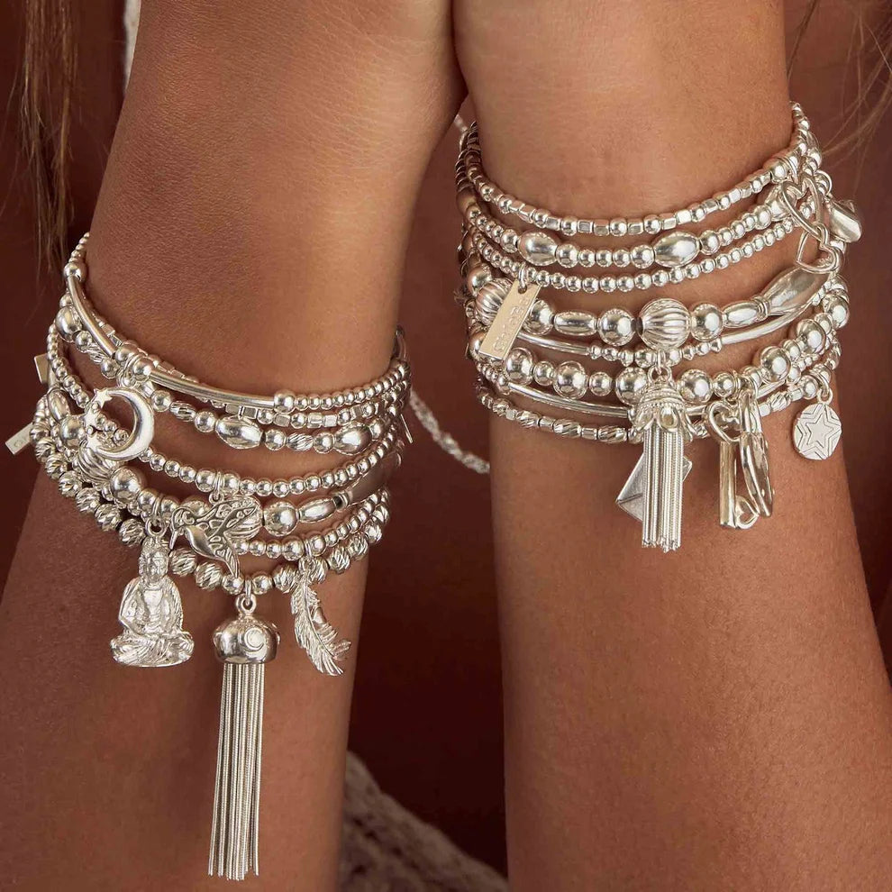 Two forearms adorned with silver beaded charm bracelets, including the ChloBo Cute Charm Hummingbird Bracelet and other positive jewelry featuring tassels, a buddha, feathers, and decorative pendants.