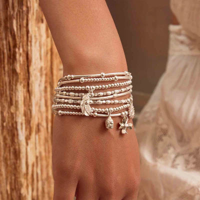 A close-up of a wrist adorned with multiple silver bracelets, including the ChloBo Cute Charm Heart In Feather Bracelet, featuring delicate beads and charms, set against a blurred background.