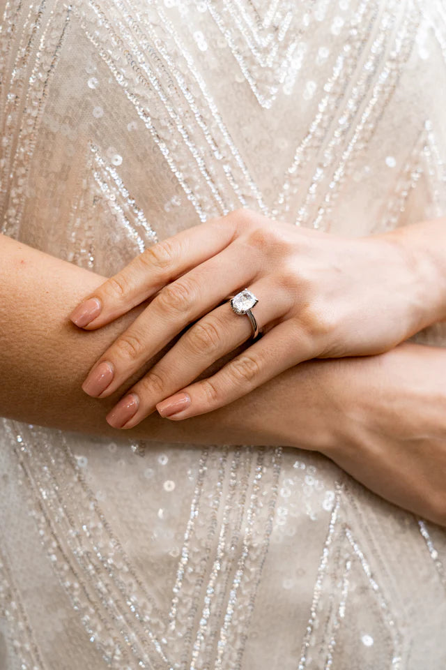 A person with manicured nails rests their hands on their arm, wearing the Georgini Sydney Soirée Elaine Cushion Cut Ring Silver on their ring finger against a background of sequined fabric.