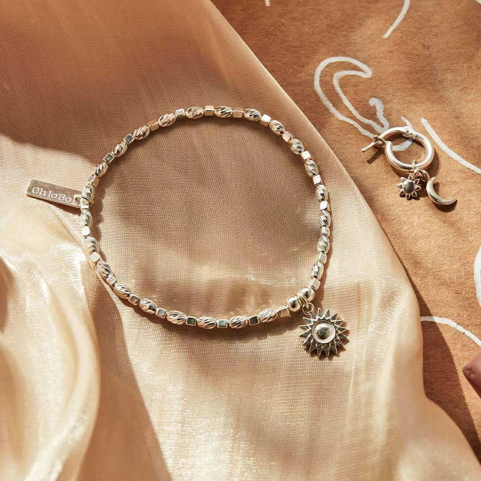 The ChloBo Soul Glow Sunshine Bracelet, a silver beaded piece with a sun-shaped charm, and hoop earrings featuring sun and moon charms, symbolizing serenity and spiritual growth, are showcased on beige fabric.