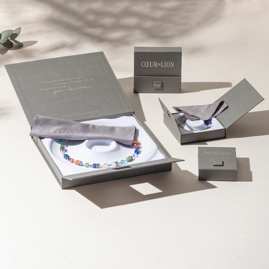 The Coeur de Lion Necklace Edge & Grace silver-gold, shown in a multicolored designer style, is displayed in an open gift box with grey cloth, next to matching earring and ring boxes on a light background.