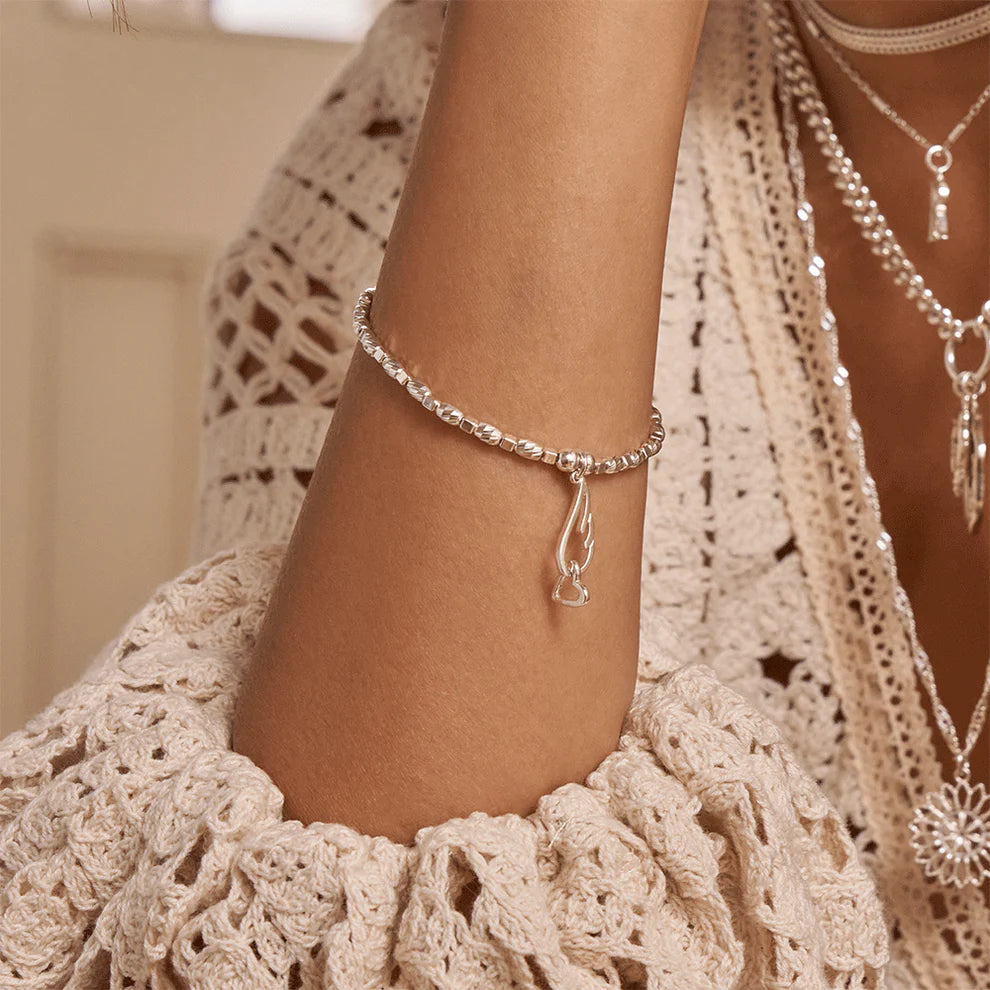 A person wears the ChloBo Soul Glow Interlocking Heart & Angel Wing Bracelet with silver beads and a pendant, paired with layered necklaces and a beige crocheted sleeve—adding meaningful, elegant style to their look.
