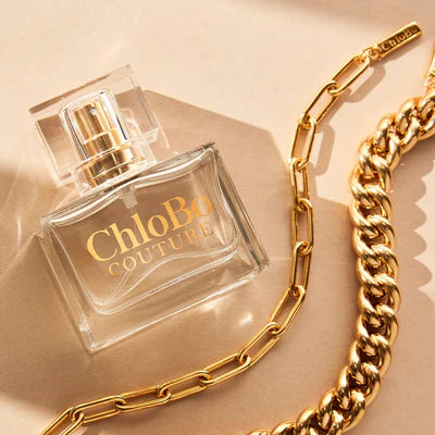 A clear 30ml bottle of ChloBo Couture Eau de Parfum sits next to two gold chain necklaces on a beige surface. This cruelty-free fragrance is displayed in elegant glass packaging.