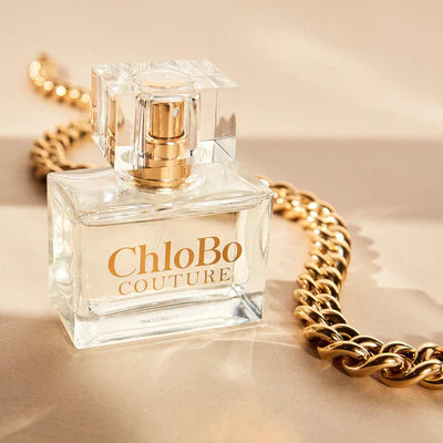 A clear glass bottle of ChloBo Couture Eau de Parfum | 30ml with a gold spray nozzle rests beside a thick gold chain on a beige surface, emphasizing this vegan and cruelty-free fragrance's elegant appeal.