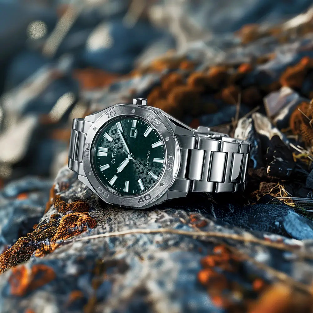 The Citizen Men's Steel Green Dial Bracelet Watch BM7630-80X, featuring a silver finish and green dial, rests on rocks covered with orange and brown lichen.