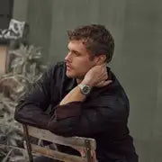 A man with short hair in a dark shirt sits sideways on a wooden chair, arms on the backrest, head turned left, showcasing the Citizen Men's Steel Green Dial Bracelet Watch BM7630-80X on his wrist.