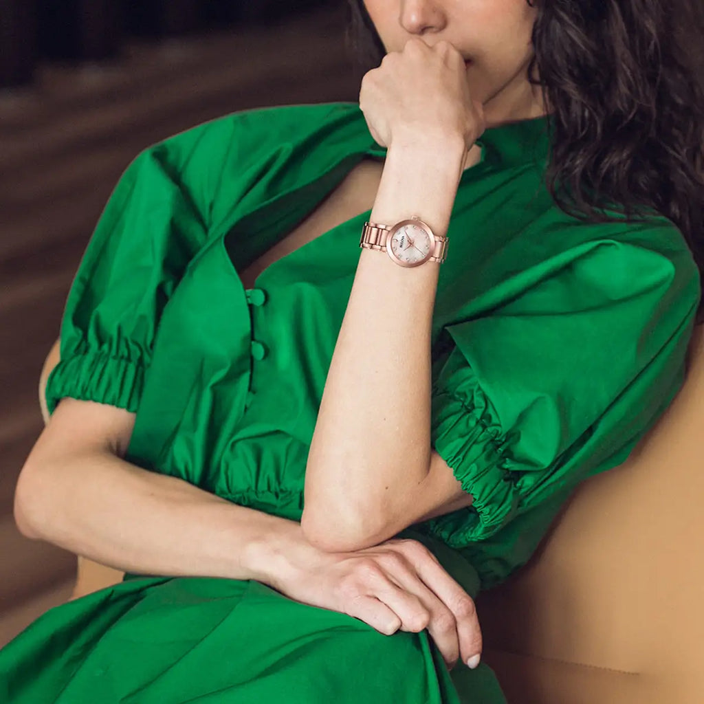A person in a green dress sits with their hand on their chin, showcasing the Bulova Ladies Futuro Diamond Watch 97P132 with a rose gold case and diamond mother-of-pearl dial on their left arm.