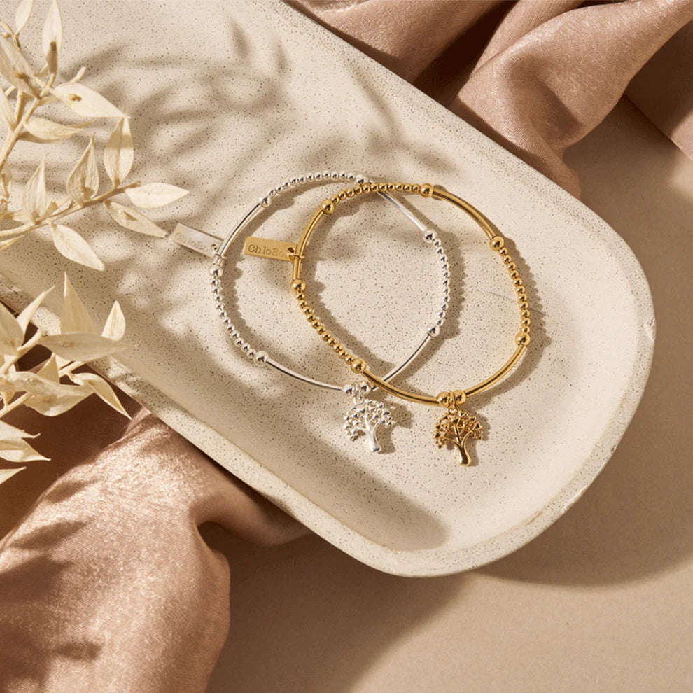 The ChloBo Cute Mini Heart Tree Of Life Bracelets, one gold and one silver with tree charms, are displayed on an oval ceramic tray with satin fabric and a dried plant—delicate symbolic jewelry perfect as gifts.