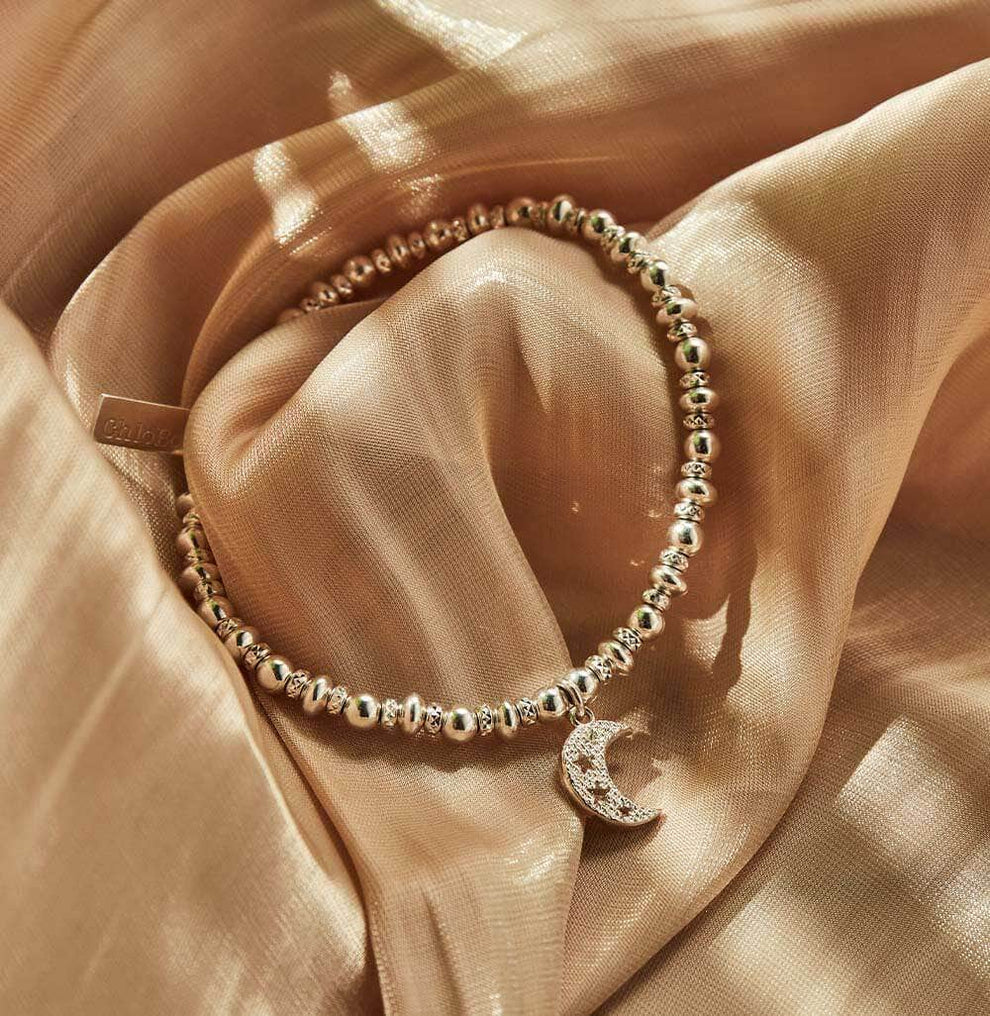 The ChloBo Didi Sparkle Starry Moon Bracelet, featuring beaded silver and a crescent moon charm, rests on shimmery light brown fabric.