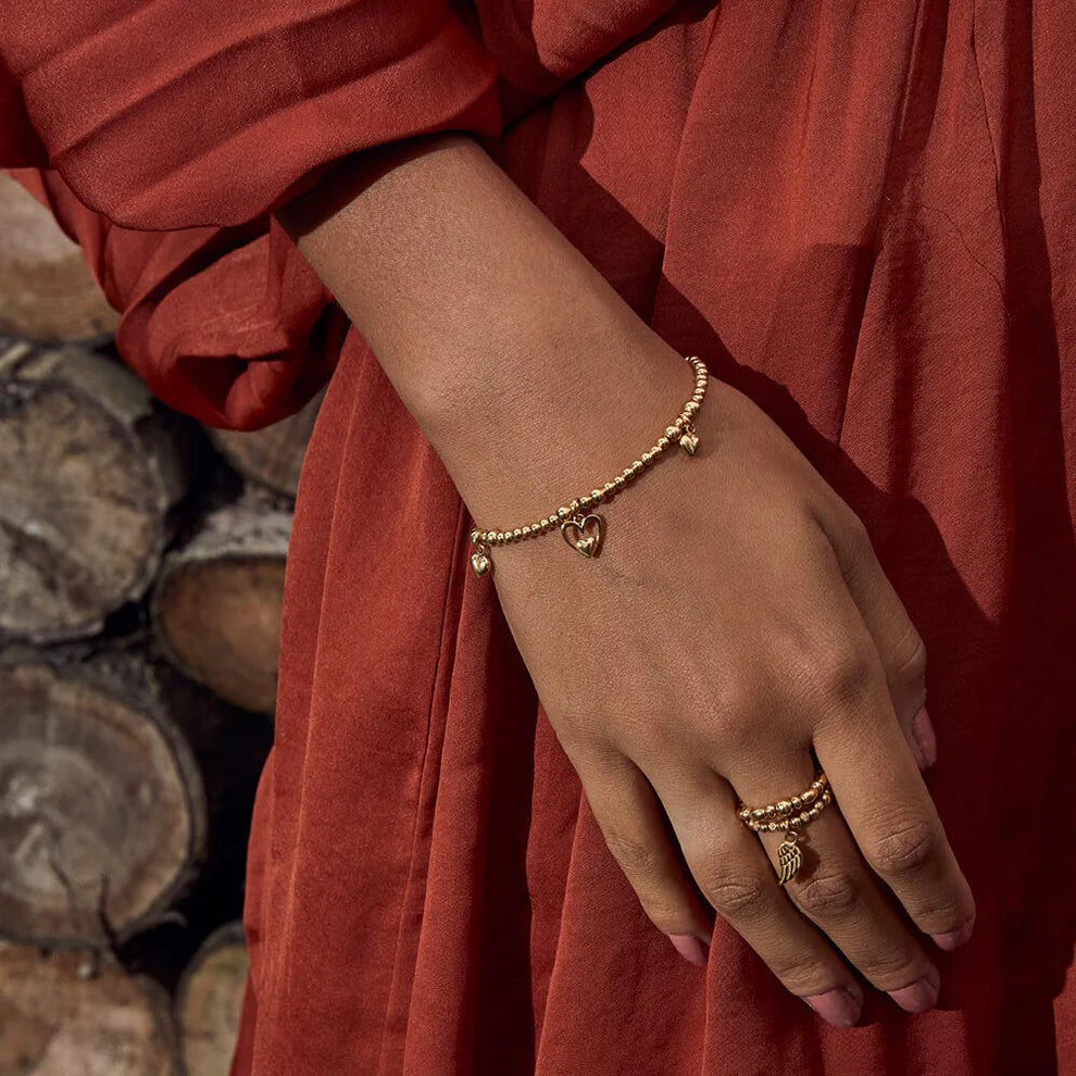 A person in a rust-colored dress wears gold beaded bracelets and rings, including the ChloBo Triple Heart Bracelet, with a stack of cut wood in the background.
