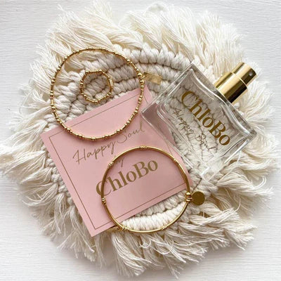Gold jewelry pieces and a bottle of ChloBo Happy Soul Eau de Parfum | 30ml are arranged on a textured white mat beside a pink card labeled "ChloBo Happy Soul Eau de Parfum.