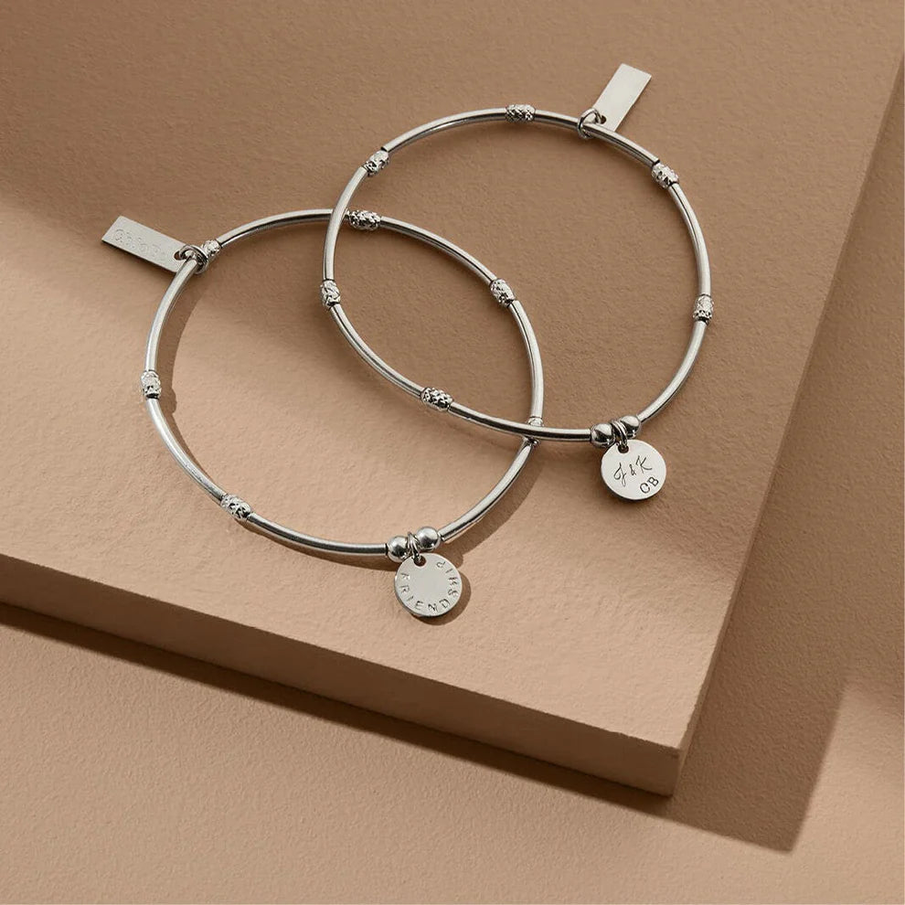 Two ChloBo Personalised Mini Noodle Sparkle Rice Friendship Disc Bracelets with round pendants are displayed on a beige geometric surface, making an ideal personalized gift.
