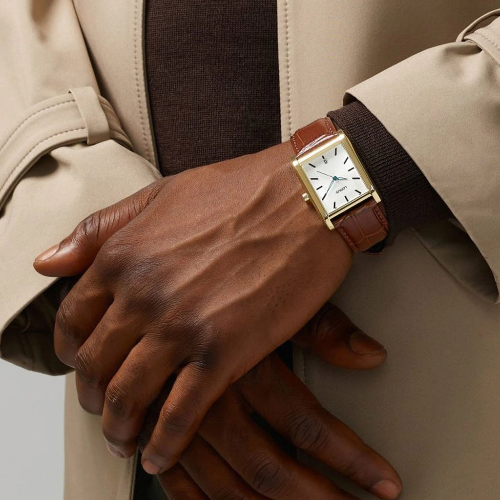 A person in a tan coat and brown sweater, hands crossed, displays the Lorus Gents White Dial Rectangle Watch RH940RX9 with a quartz movement and brown leather strap.