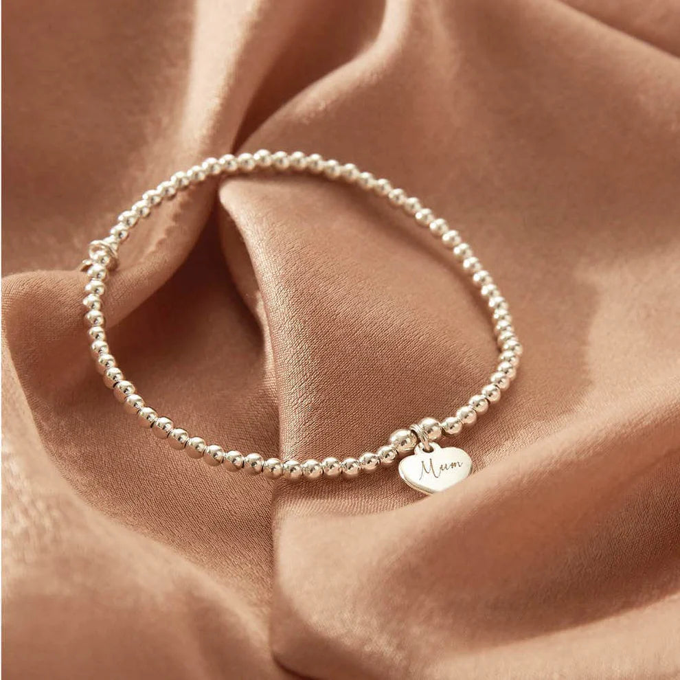 The ChloBo Personalised Cute Charm Heart Bracelet, featuring silver beads and a small "Mum" engraved oval charm, is displayed on beige satin fabric.