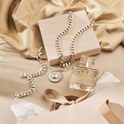 A bottle of ChloBo Couture Eau de Parfum | 30ml, two silver chain bracelets, and a pendant rest on beige satin with a gift box and ribbon—a chic showcase of elegant jewelry and cruelty-free fragrance.