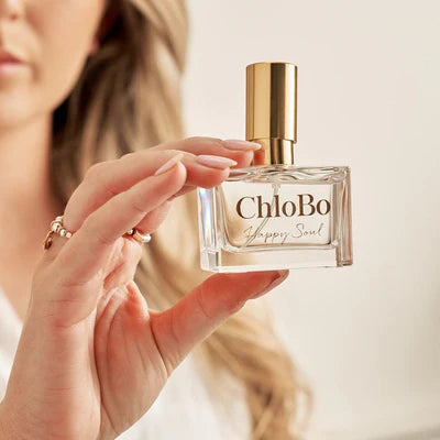 A woman holds a clear glass bottle of ChloBo Happy Soul Eau de Parfum | 30ml with a gleaming gold cap, displaying its elegant vegan label to the camera.